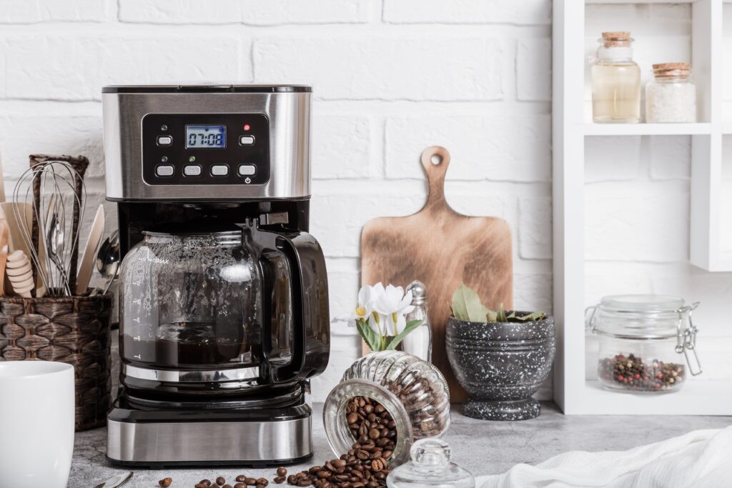 A black drip electric coffee machine with a glass teapot brews a A coffee maker on the counter next to spilled coffee beans.