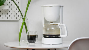 A small white coffee maker on a table next to a cup of coffee.