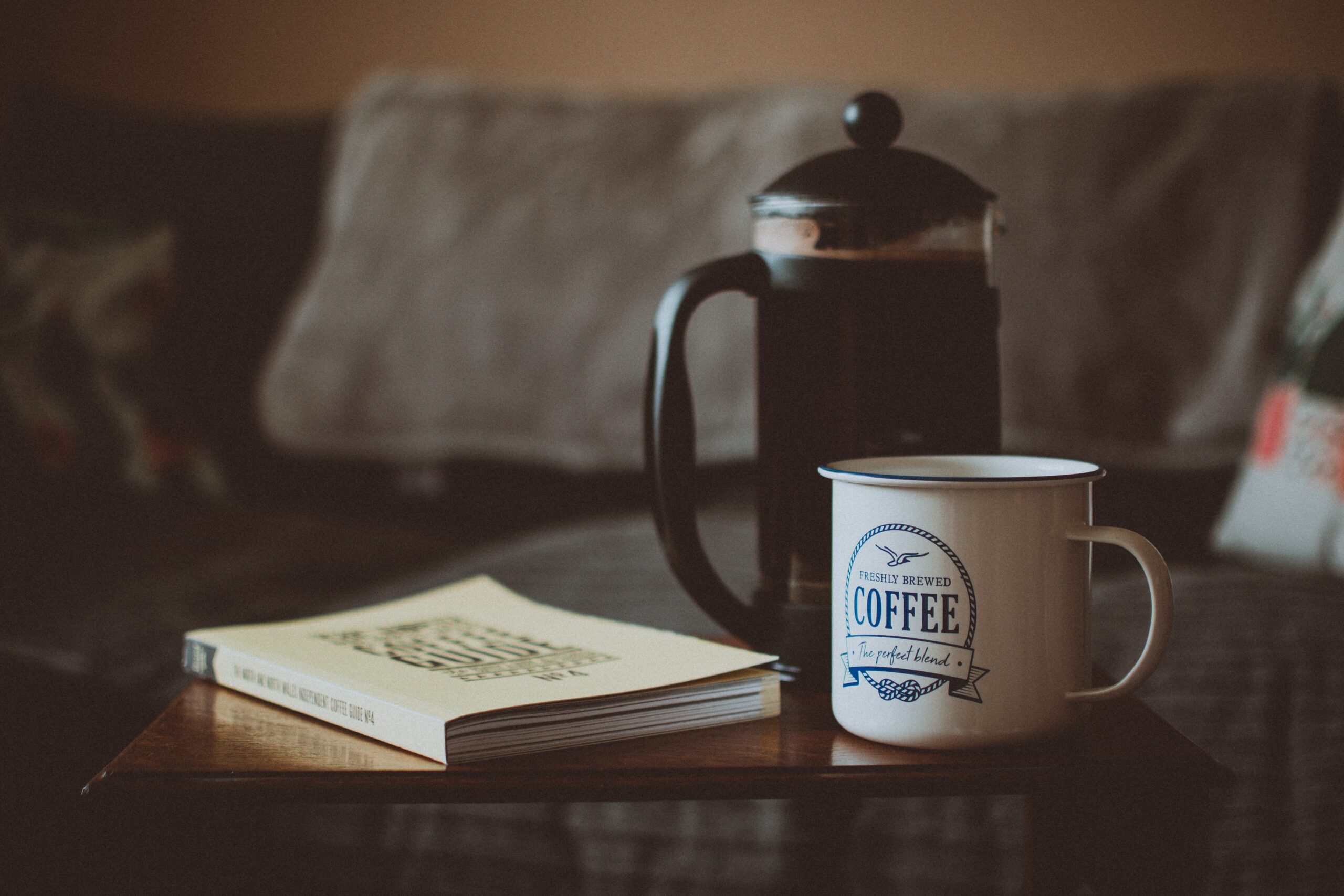 A French press on a table next to a coffee mug and a book.